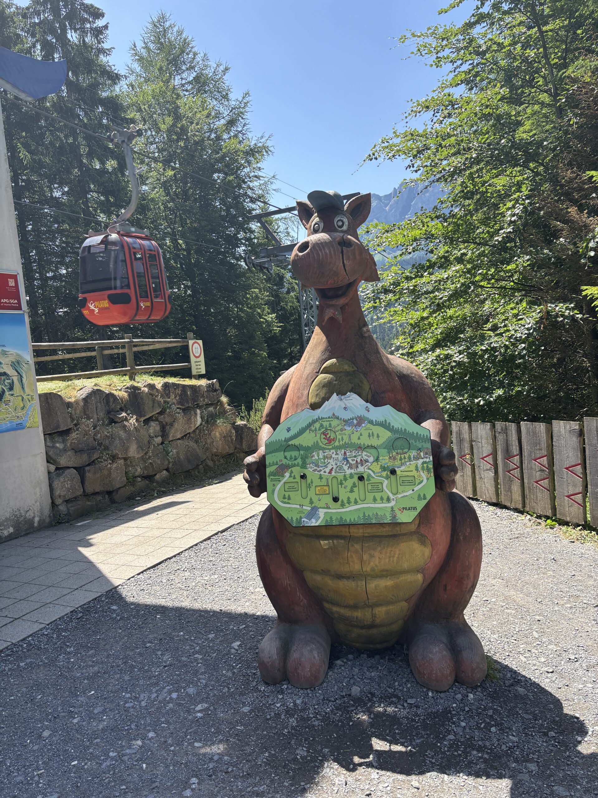 Große Drachenfigur bei Fräkmüntegg am Pilatus mit Übersichtskarte der Drachenwelt und vorbeifahrender roter Pilatus-Gondelbahn.